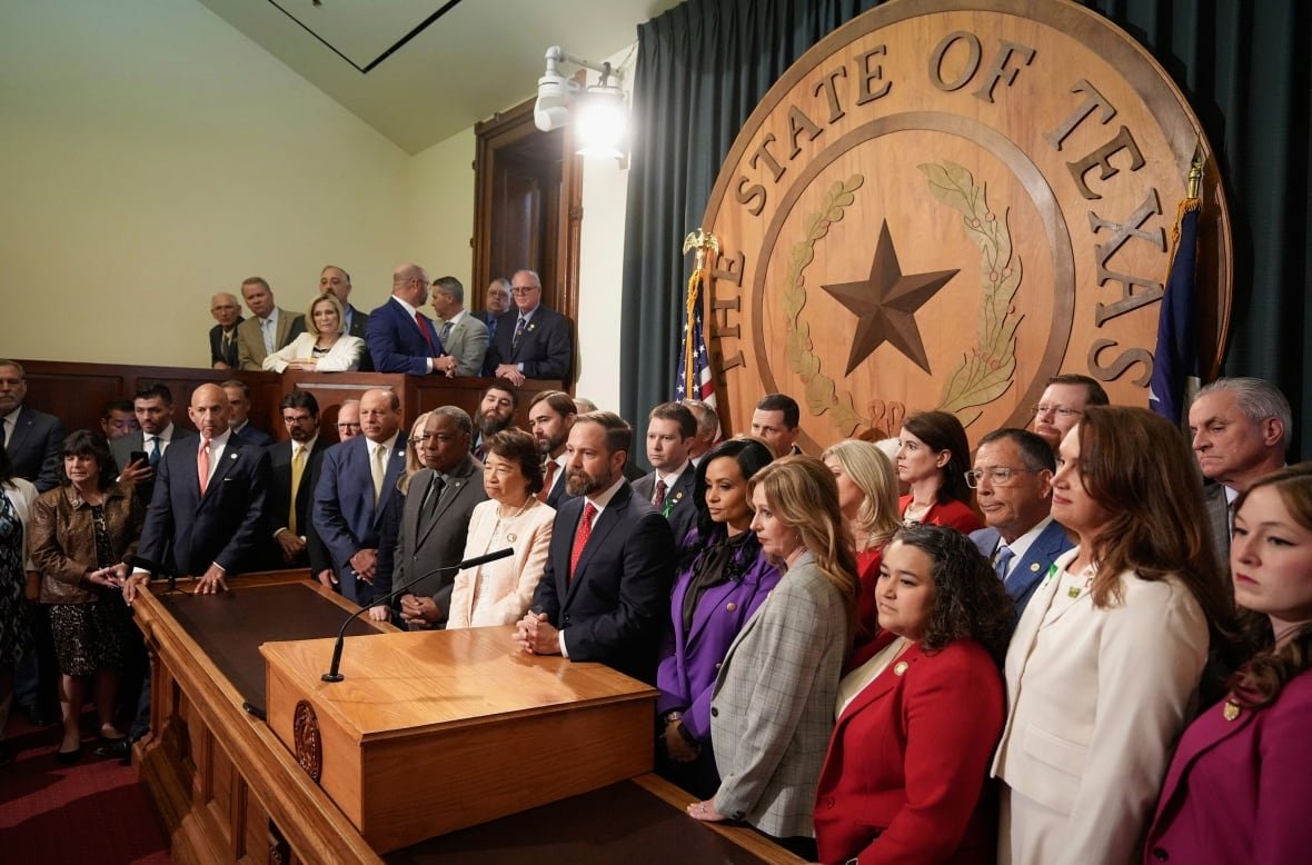 A large group of people standing behind a podium in a legislature building in front of a large wooden sign that says "The State of Texas."