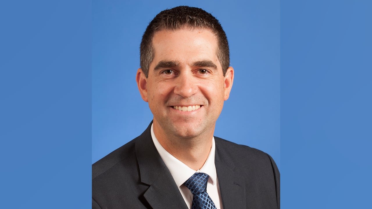 default-632 A portrait of a smiling man in a dark suit against a blue background