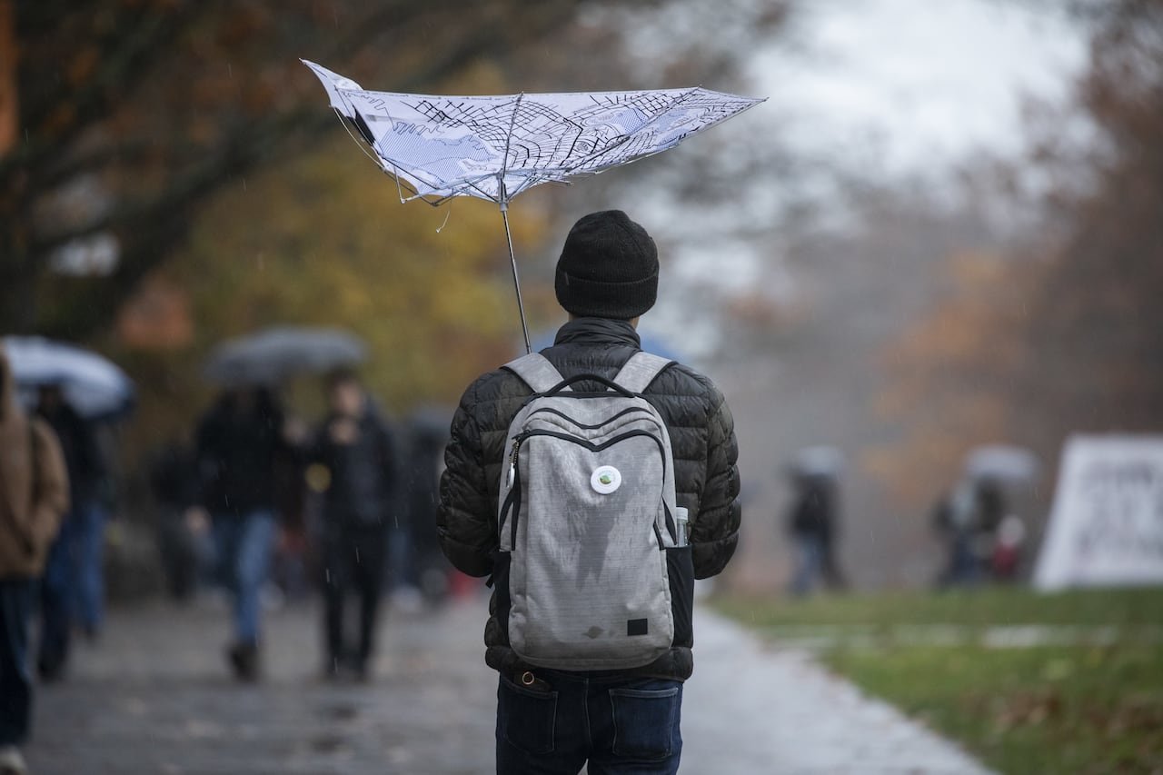 A student is shown wearing a backpack and holding an umbrella that has been flipped inside out by the wind.